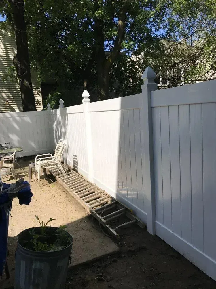 White vinyl fence beside a narrow dirt path with stacked ladders and a chair in a shaded backyard