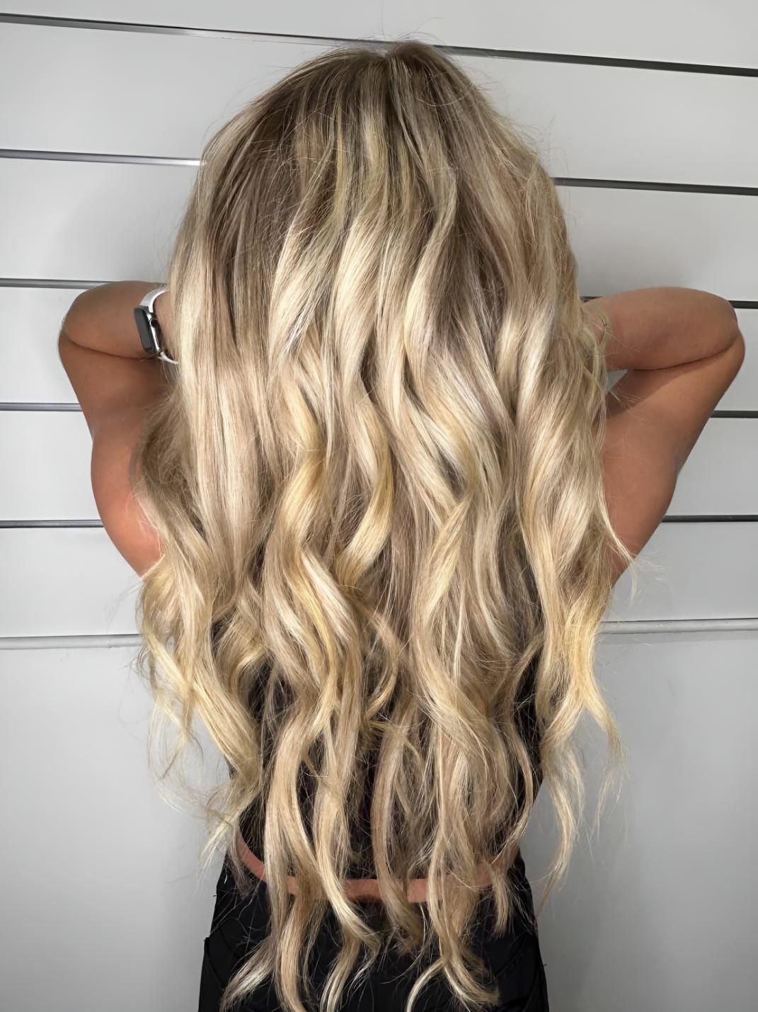 A Woman With Long Blonde Wavy Hair is Standing in Front of a White Wall — Strut Hair & Beauty In Maroochydore, QLD