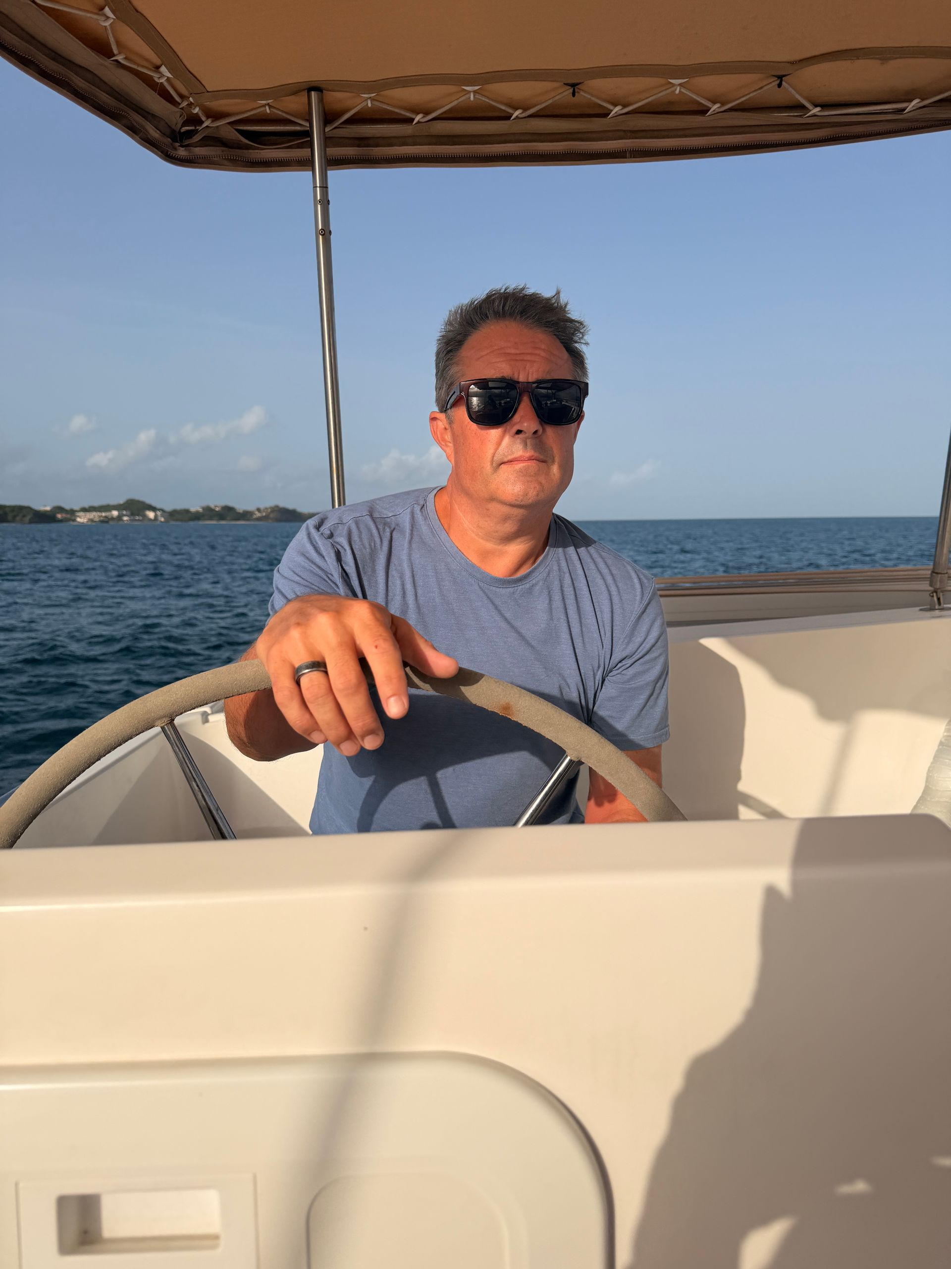 Sexy man steering a boat on calm water, wearing sunglasses and a blue shirt.