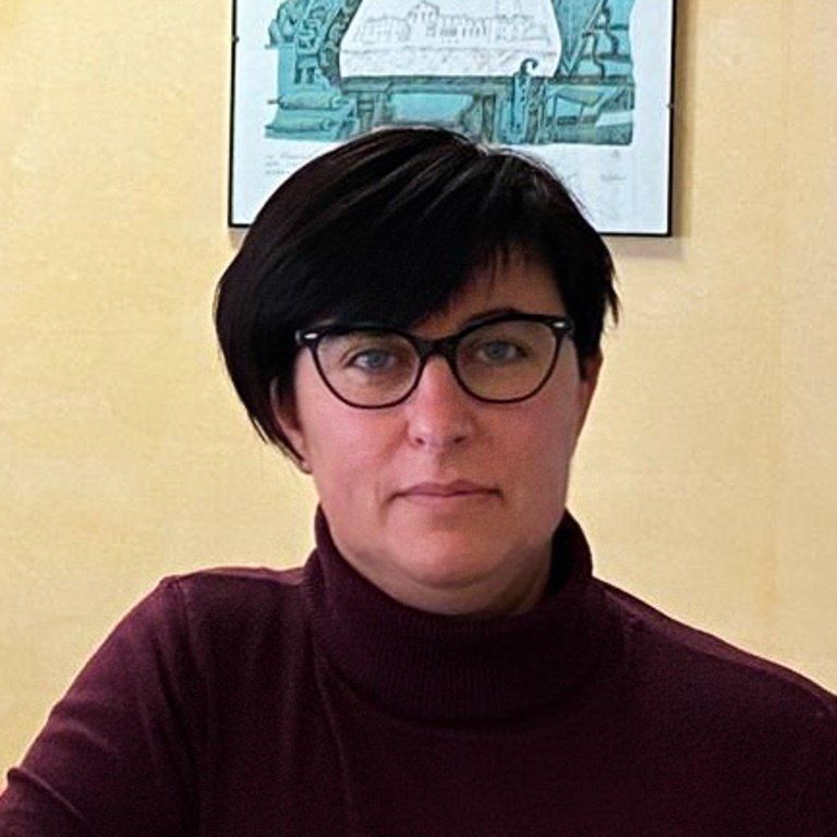 Una donna con gli occhiali e un dolcevita è seduta davanti a un quadro sul muro.