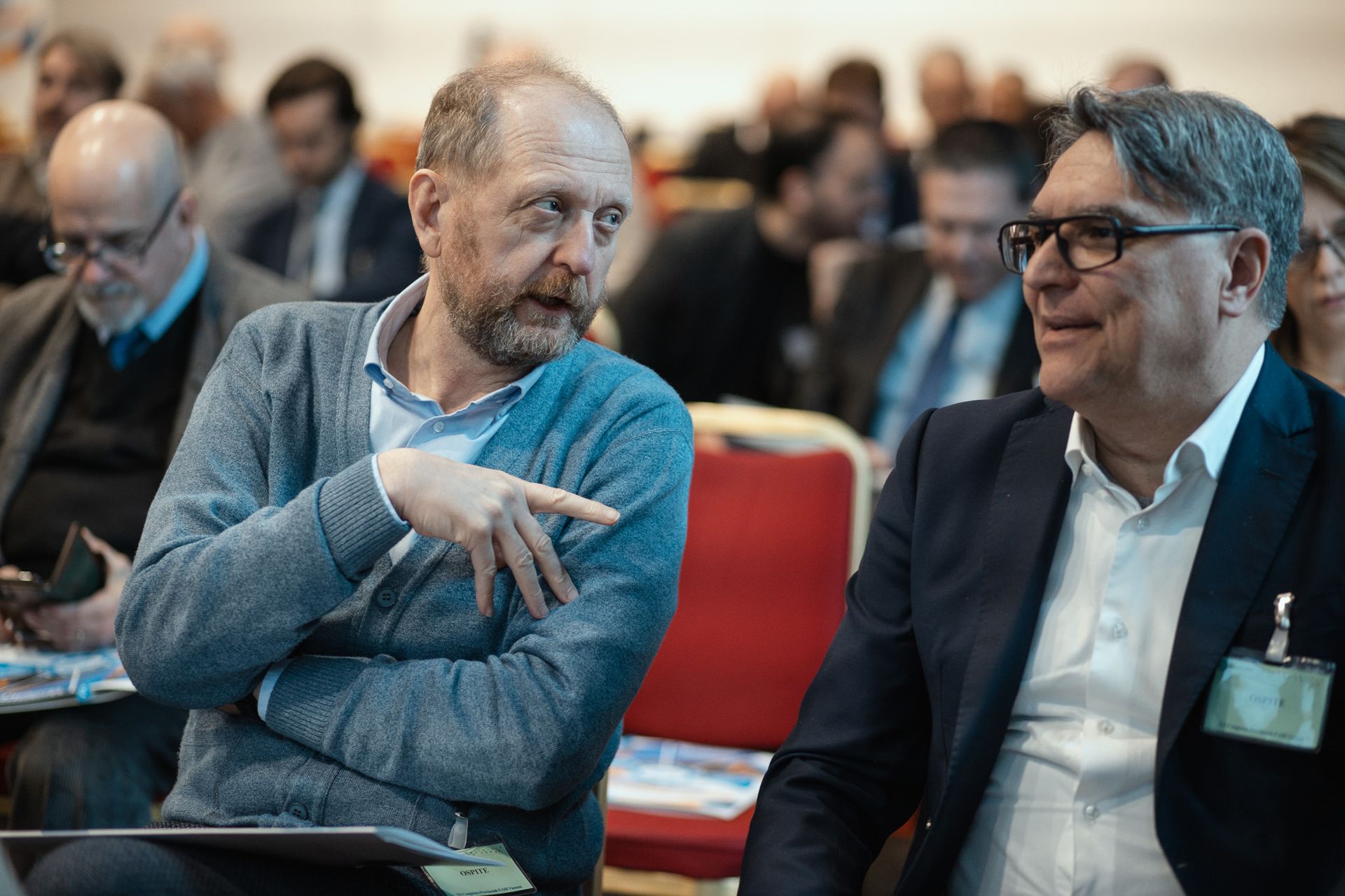 Due uomini sono seduti in una sala conferenze e parlano tra loro.