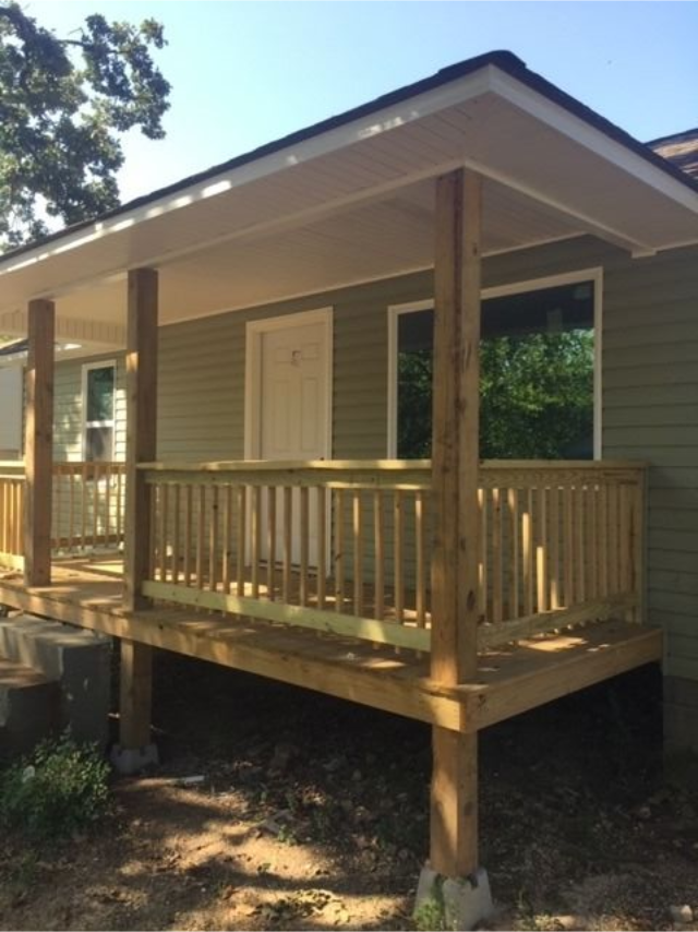 Covered wooden porch on a small house with railings and steps, in a wooded yard.