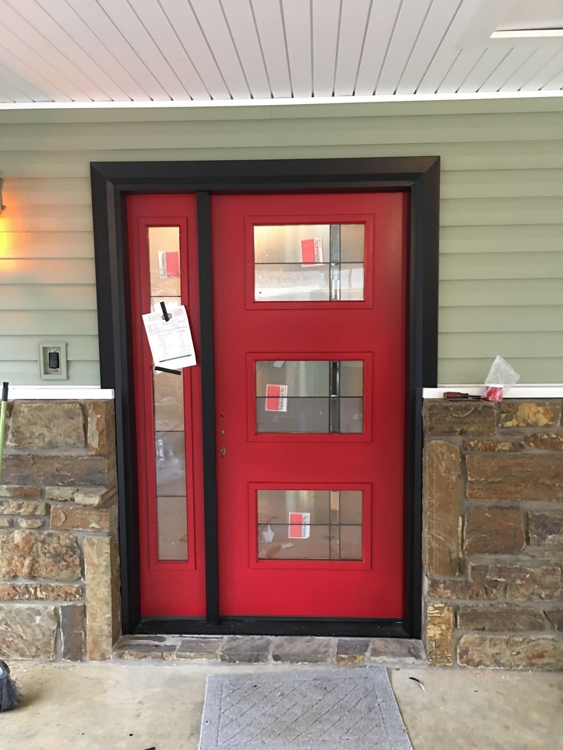 Red front door with three windows and a notice, framed by stone walls under a porch ceiling