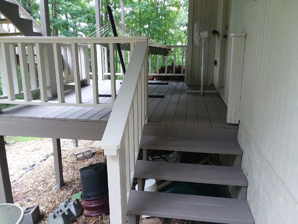 Covered porch with wooden steps, railing, and white siding beside a small outdoor deck.