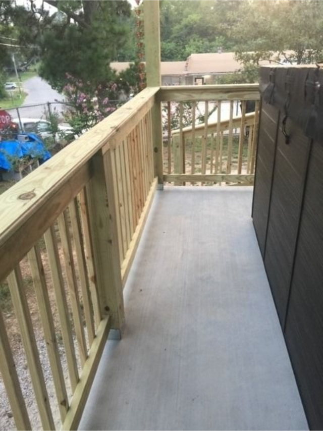 Narrow wooden balcony with railing, concrete floor, and trees visible beyond the deck.