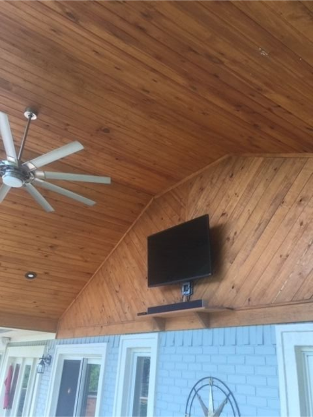 Covered patio with wood ceiling, ceiling fan, wall-mounted TV, and light blue brick wall.