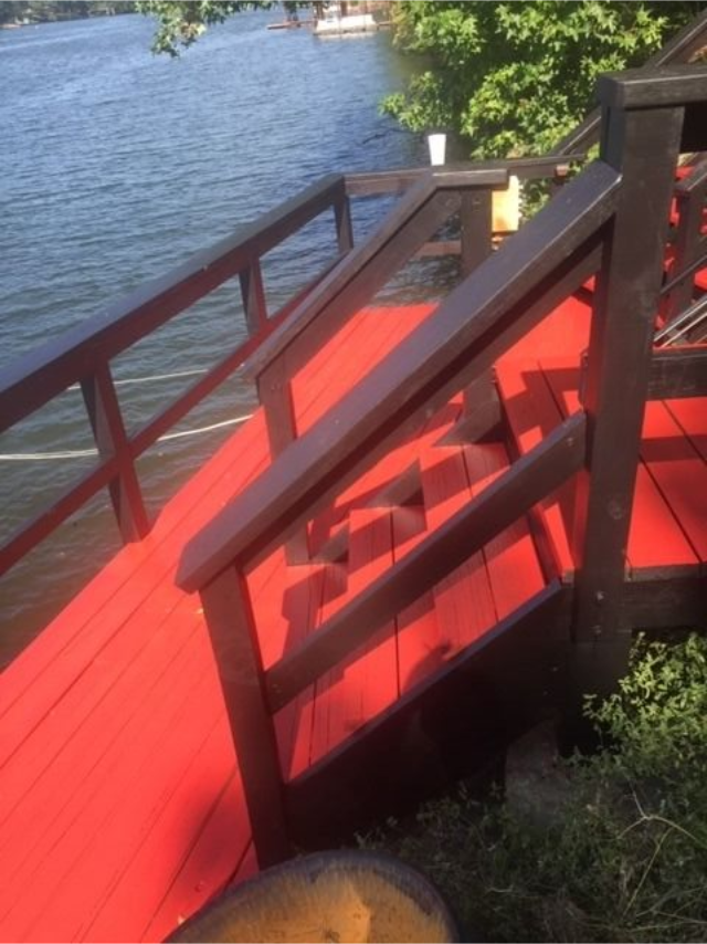 Red wooden stairway with railing leading down to a dock beside a calm lake under trees