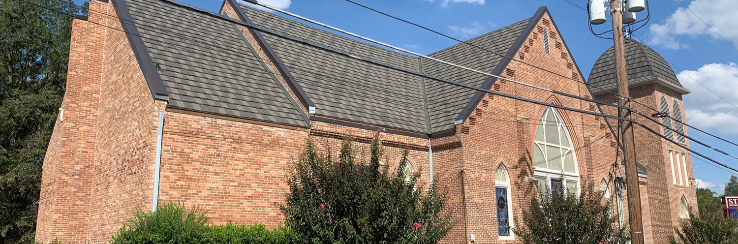 Brick church building with a gray shingled roof and arched window under a blue sky