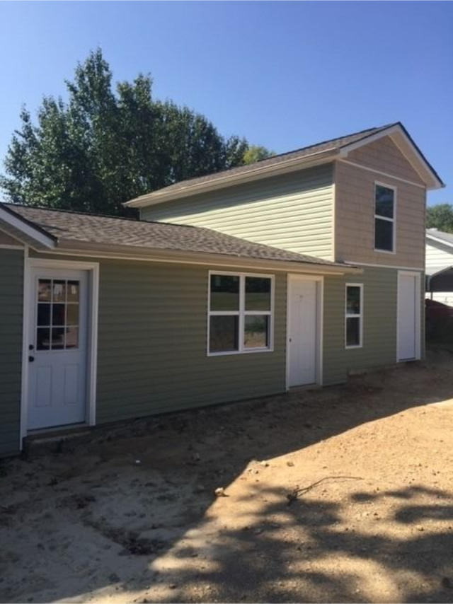 Small green house exterior with white trim, a side door, and a two-story section under construction.