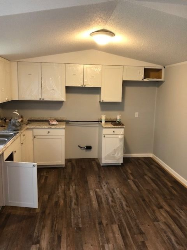 Empty kitchen with white cabinets, gray walls, and dark wood flooring under a ceiling light.