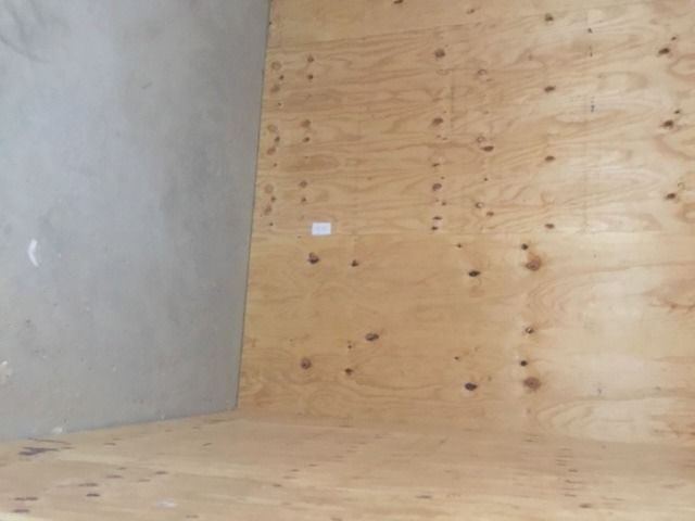 Bare plywood wall and floor in an unfinished room with a small white label on the wall.