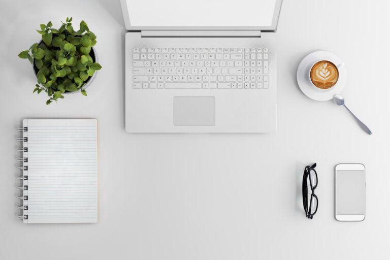 A white desk with a laptop, cell phone, glasses, and a cup of coffee.
