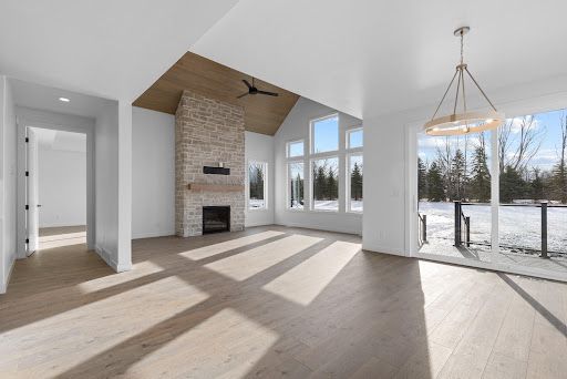 The great room, dining area and outdoor views come together in one light-filled living space. Bright empty living room with stone fireplace, wood floor, and large windows overlooking snowy trees