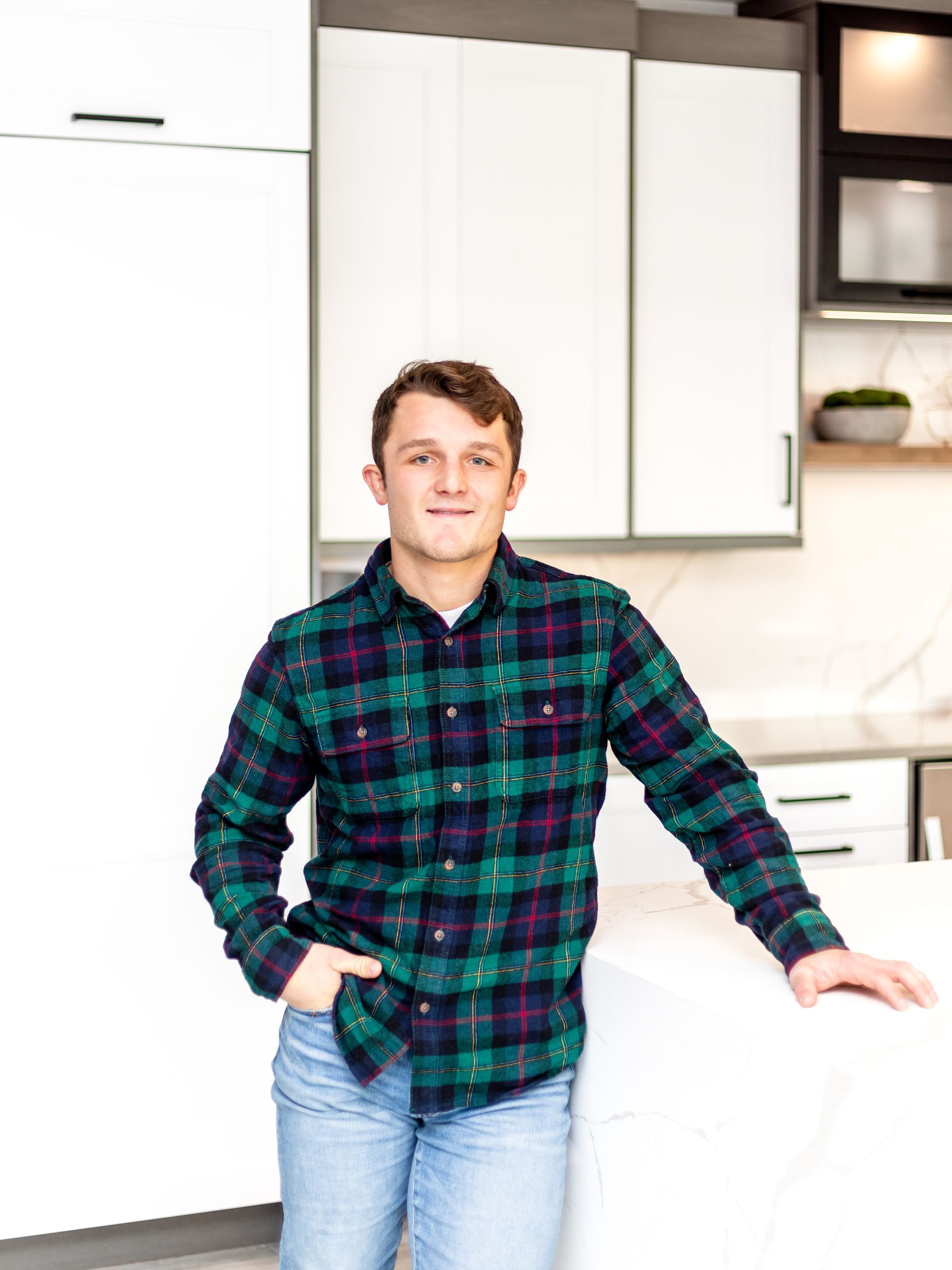 Man in plaid shirt and jeans, leaning on a white kitchen island, smiling, modern kitchen in background.