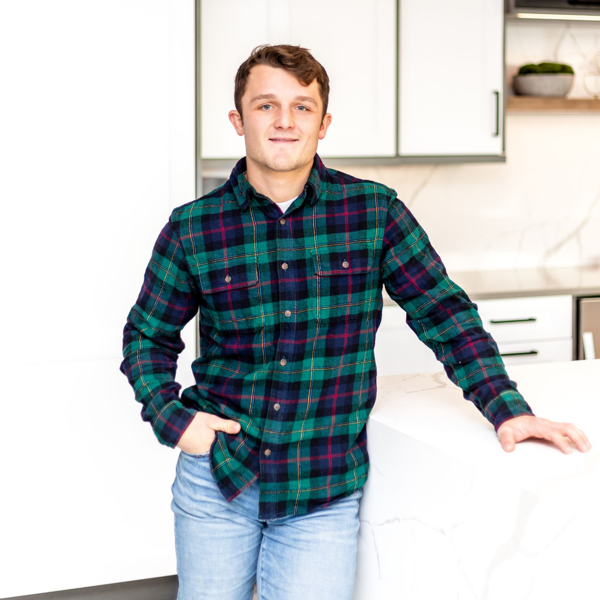 Man in plaid shirt and jeans, leaning against a white countertop in a modern kitchen.