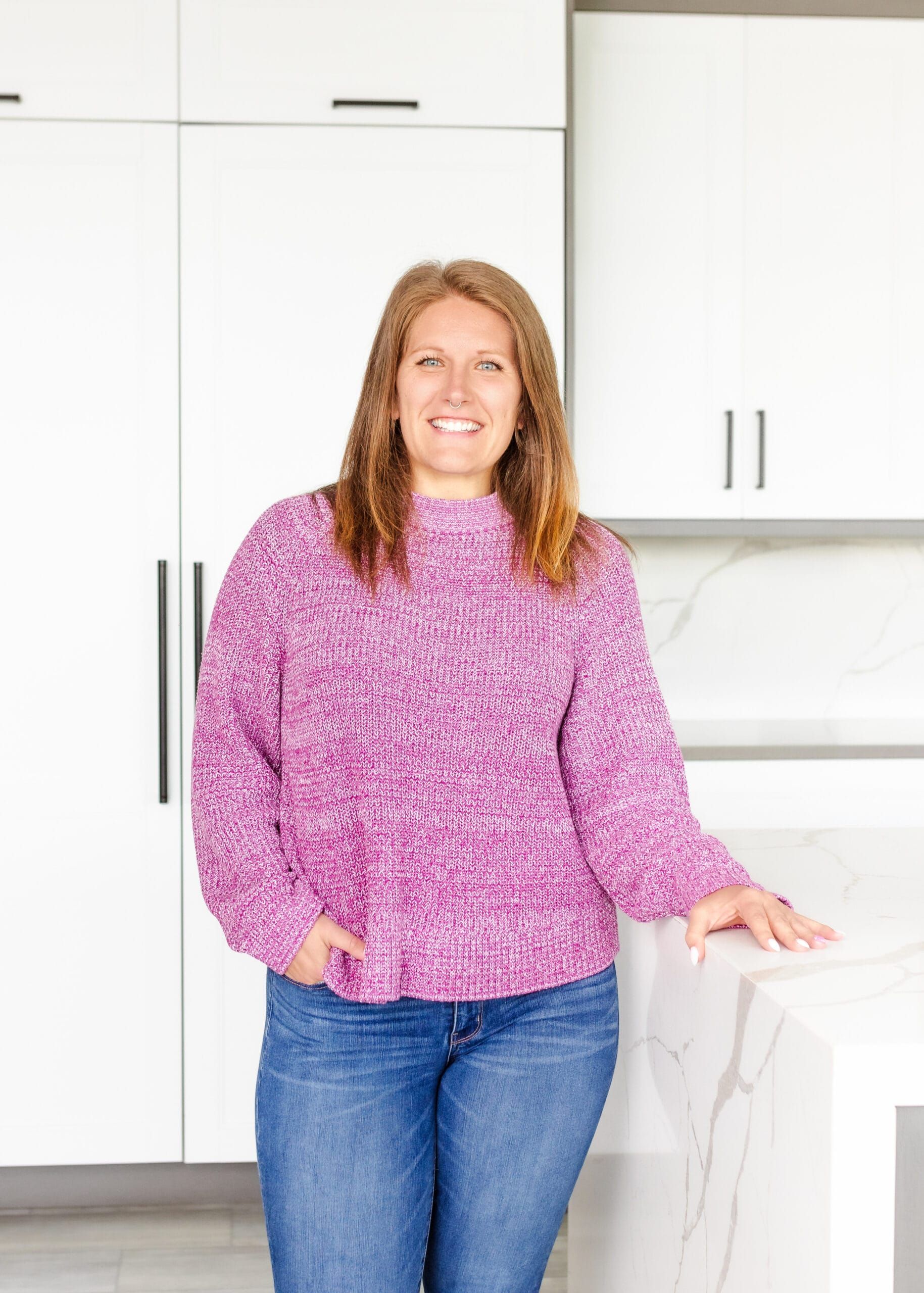 Woman in pink sweater and jeans in a modern white kitchen, smiling, leaning on a counter.