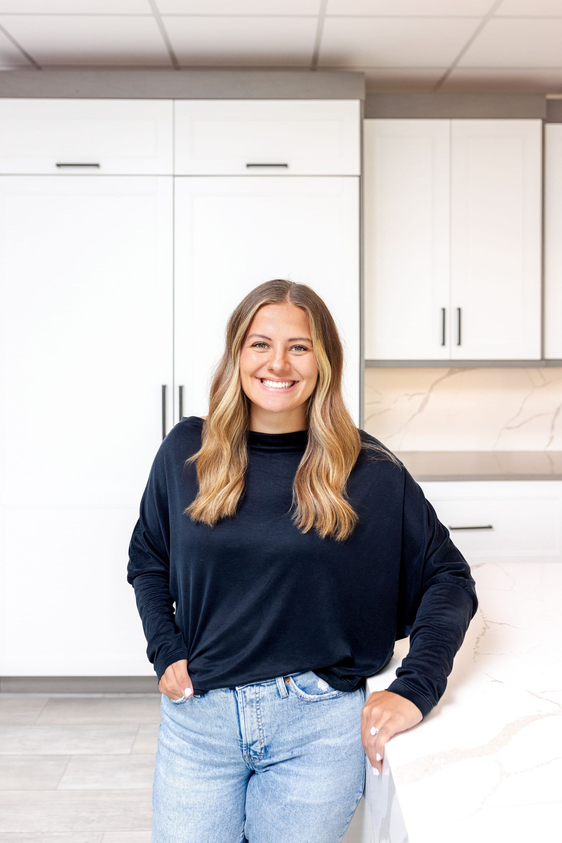 Woman smiling in a kitchen, leaning on a counter. She wears a black top and jeans. White cabinets in the background.