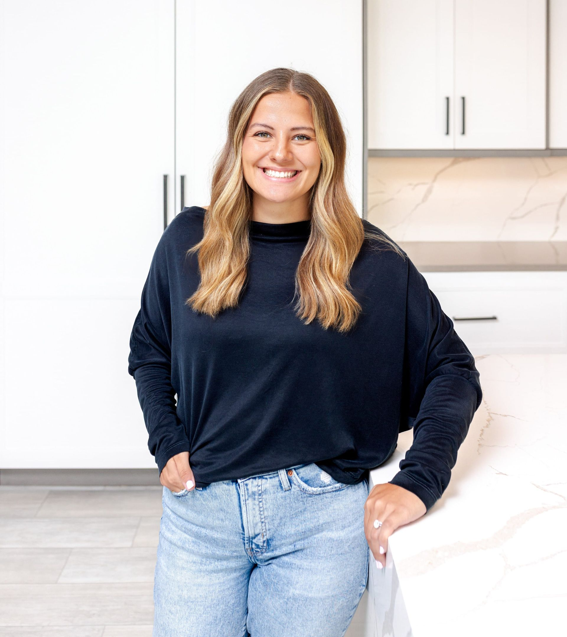 Woman in a kitchen smiling, wearing a black shirt and jeans, leaning on a white countertop.