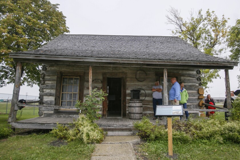 Bonanzaville Pioneer Village and Museum A rustic log cabin with a wooden porch and shake roof, featuring a small barrel and a sign standing in the grassy yard.