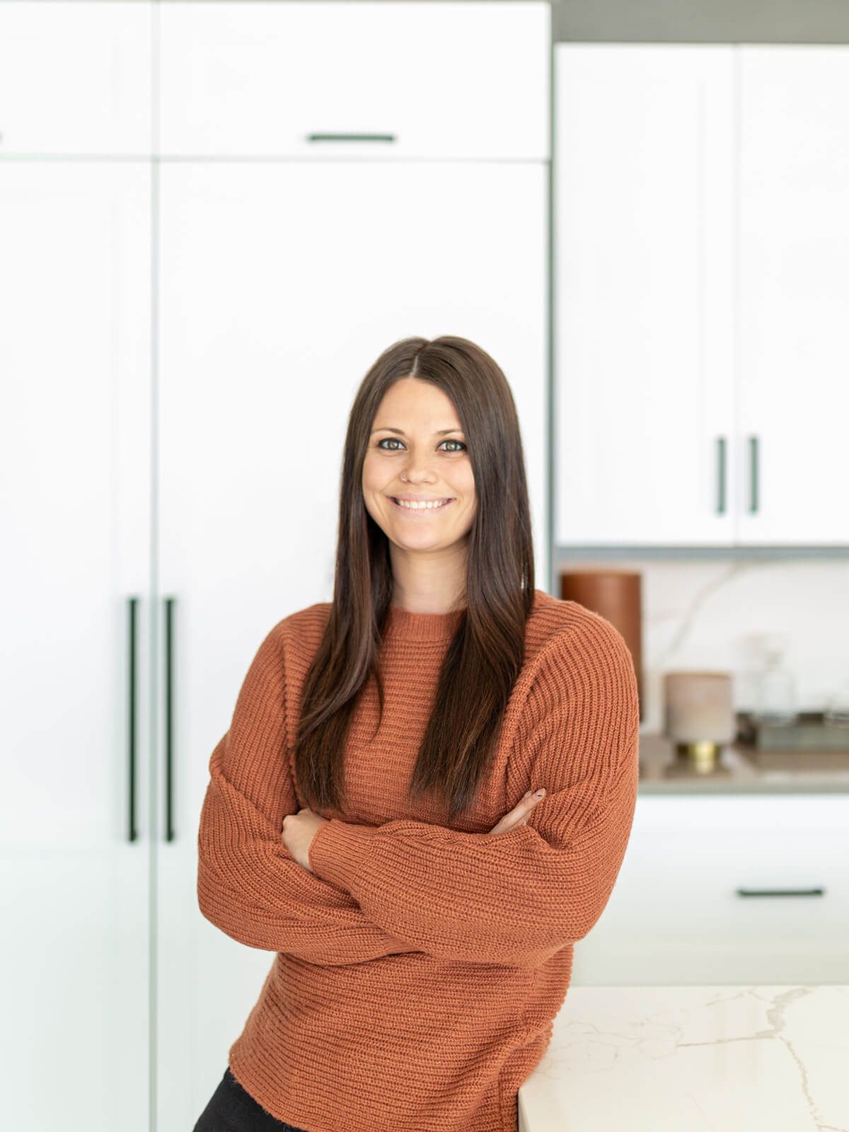 Woman in orange sweater smiles, arms crossed, in a modern kitchen with white cabinets.