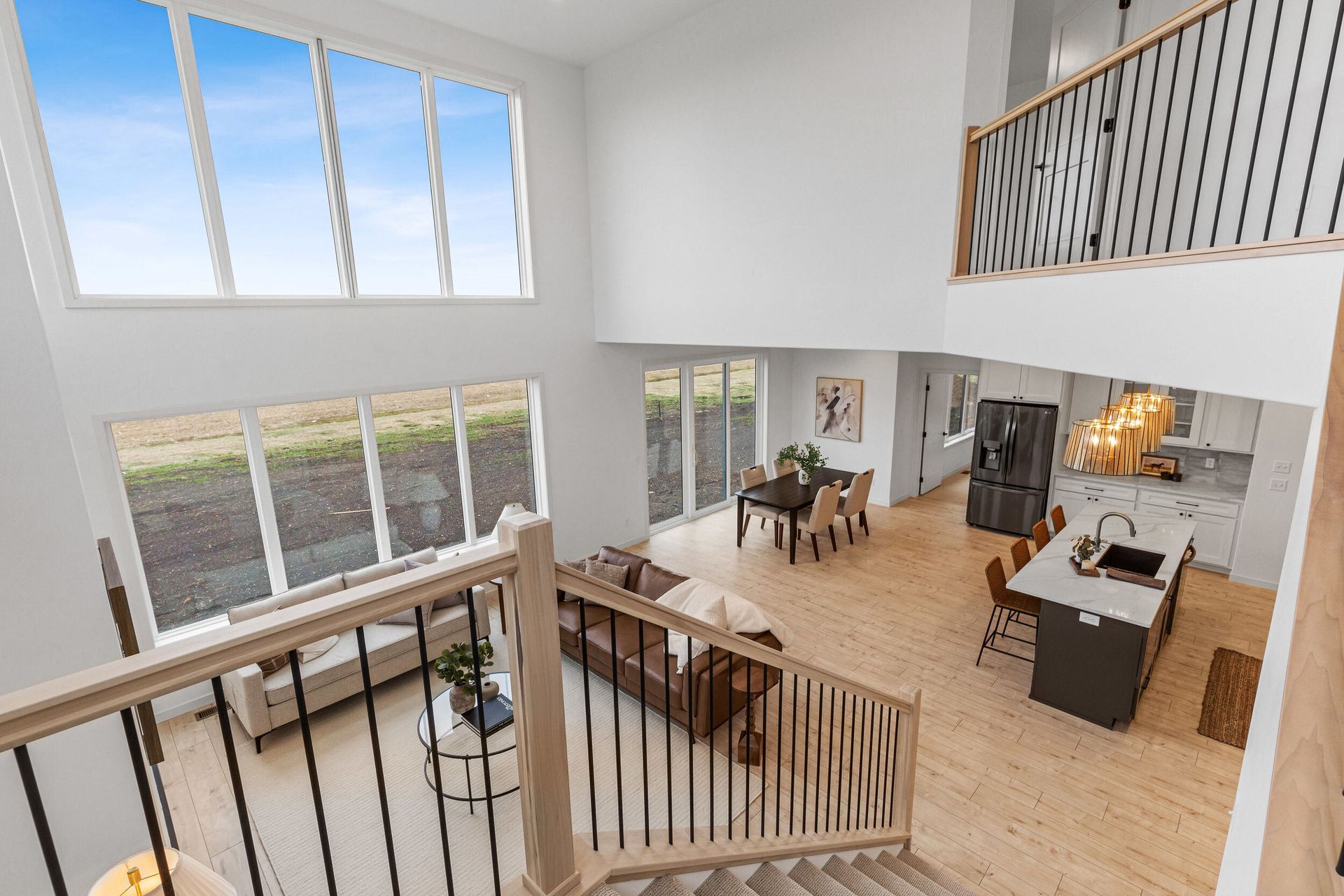 Open-concept home interior with large windows, light wood floors, and a staircase leading to a second floor balcony.