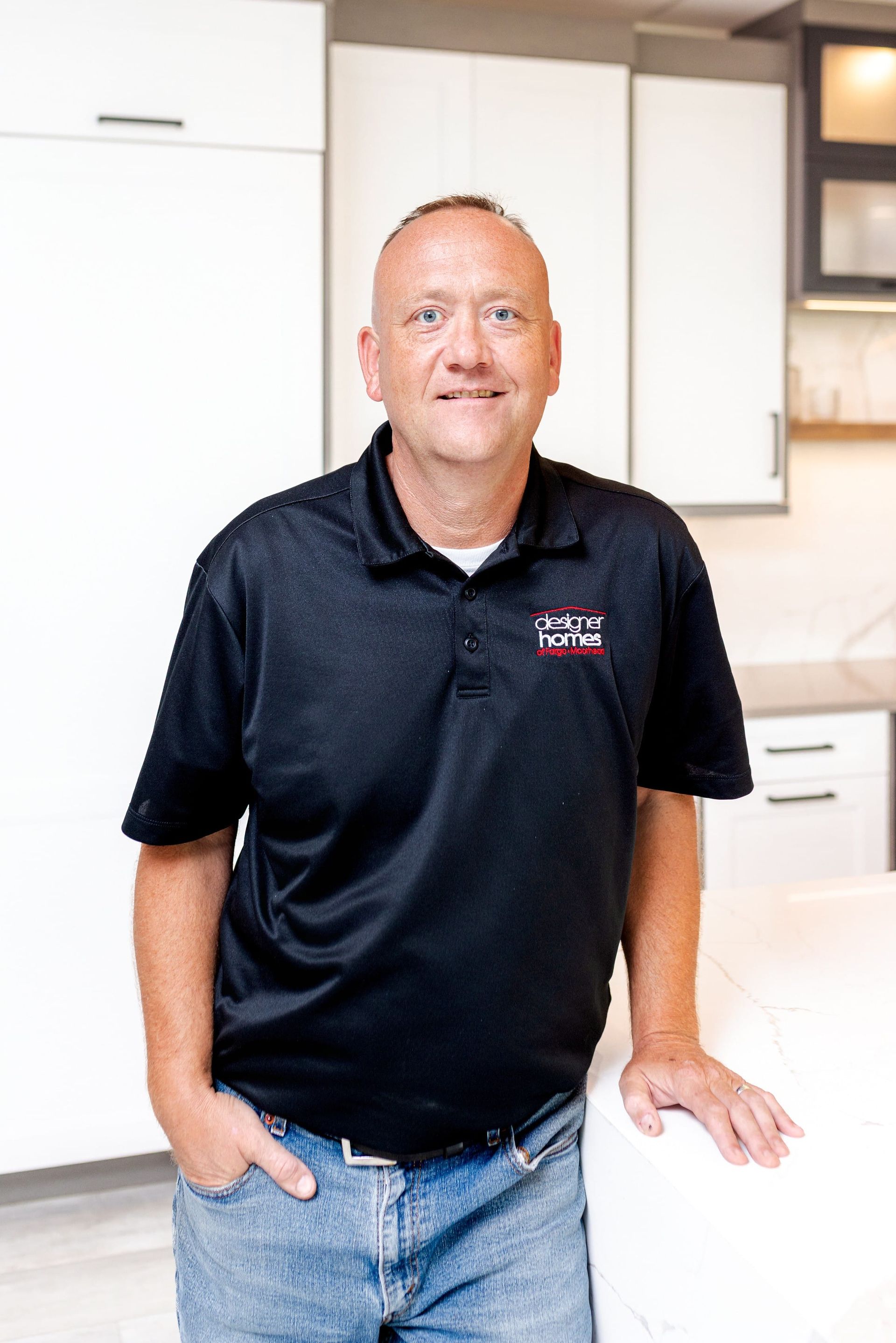 Man in black polo shirt stands in kitchen, leaning on a counter.