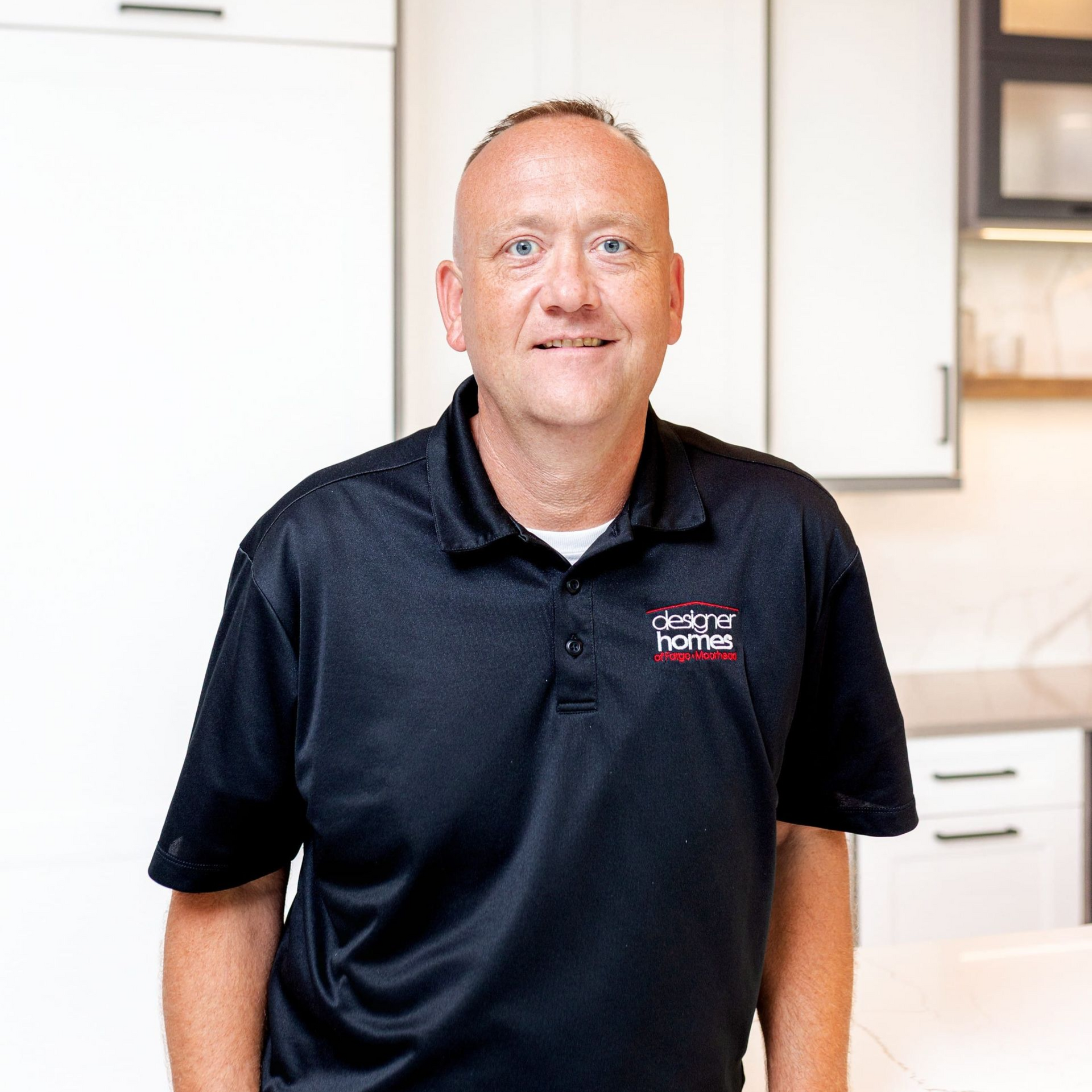 Man in black polo shirt stands in kitchen, leaning on countertop.