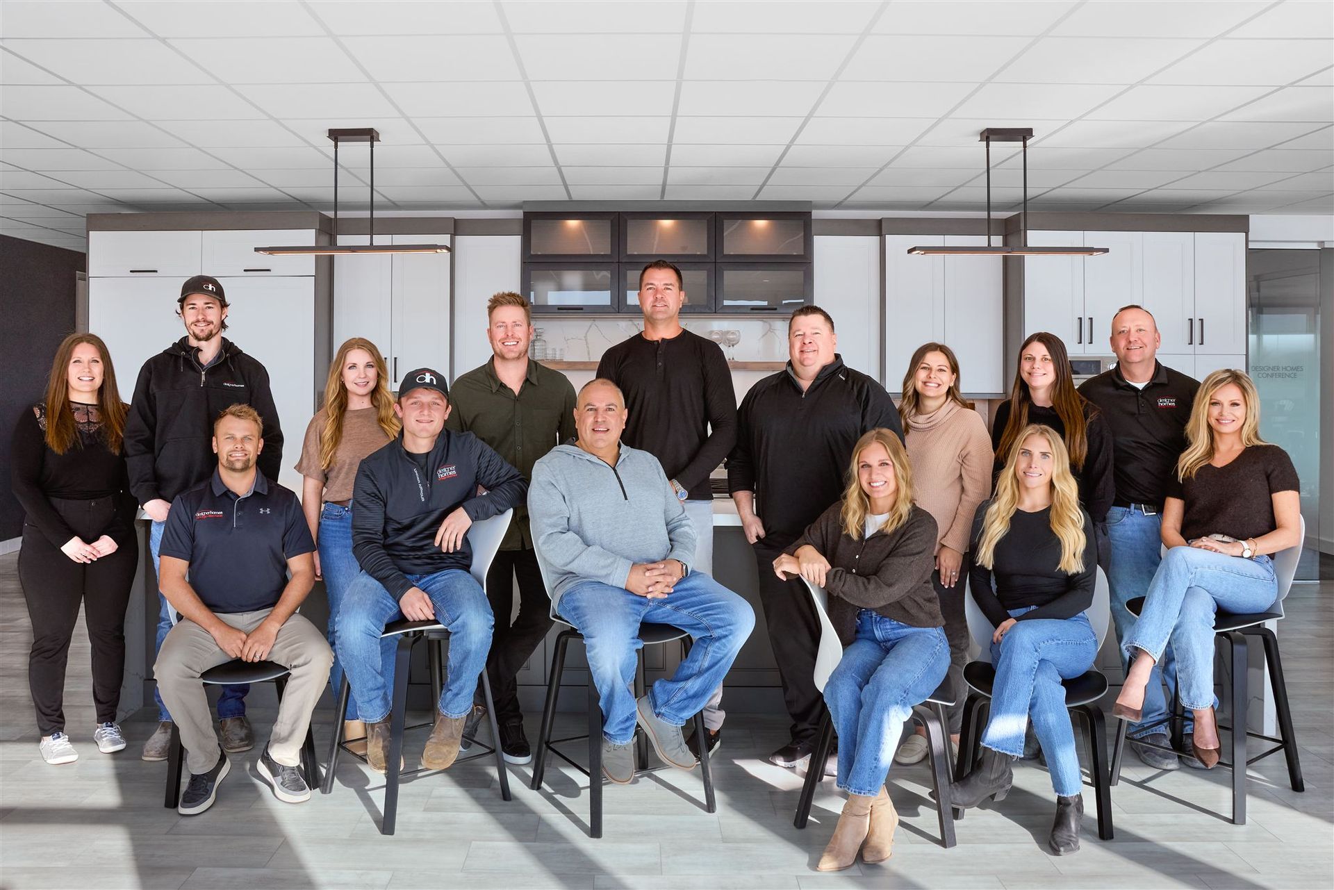 A group of fifteen people in casual business attire pose for a photograph in a modern office kitchen.