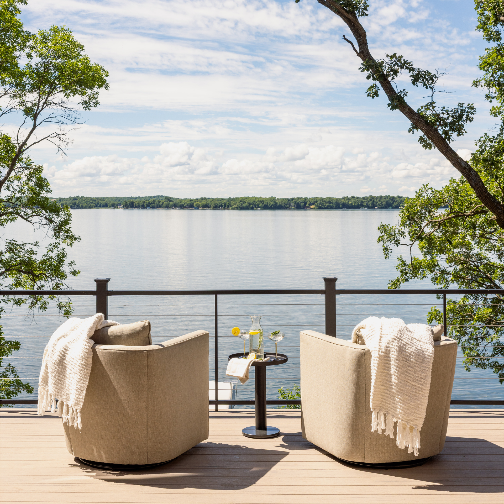 Two beige chairs on a lakefront deck with a small table and white towels, facing calm water and trees.