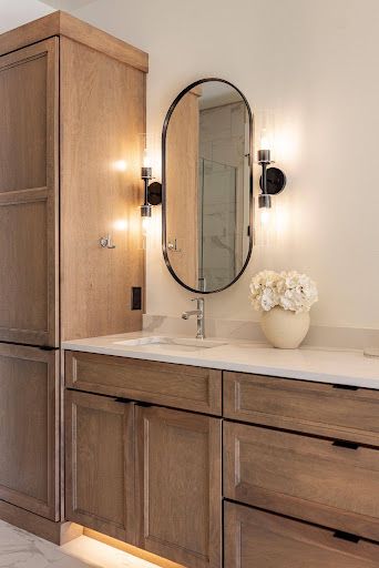 Wood vanity with oval mirror, wall sconces, and white flowers in a beige bathroom.