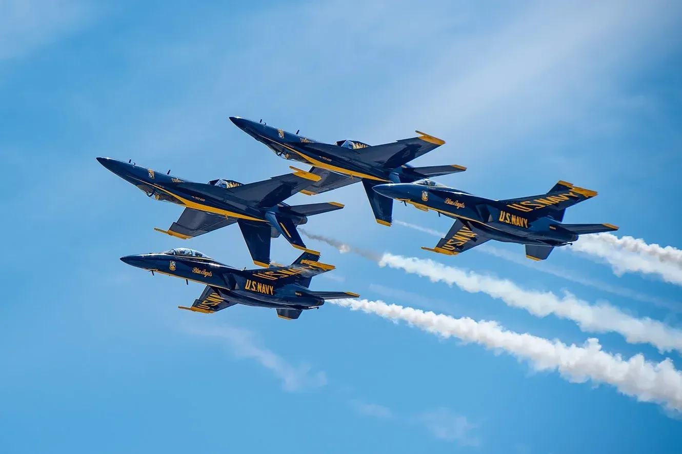 Fargo AirSho Four Blue Angels F/A-18 Hornet jets flying in a tight formation against a bright blue sky, leaving long white smoke trails.