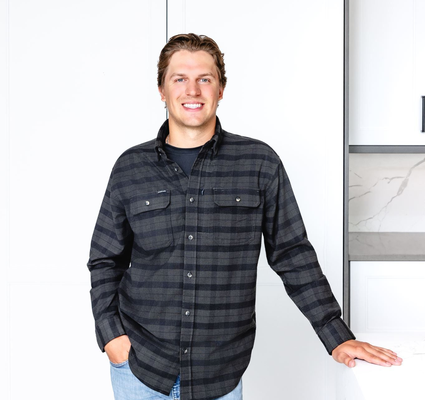 Smiling person in a dark plaid shirt standing against a white wall, one hand in pocket
