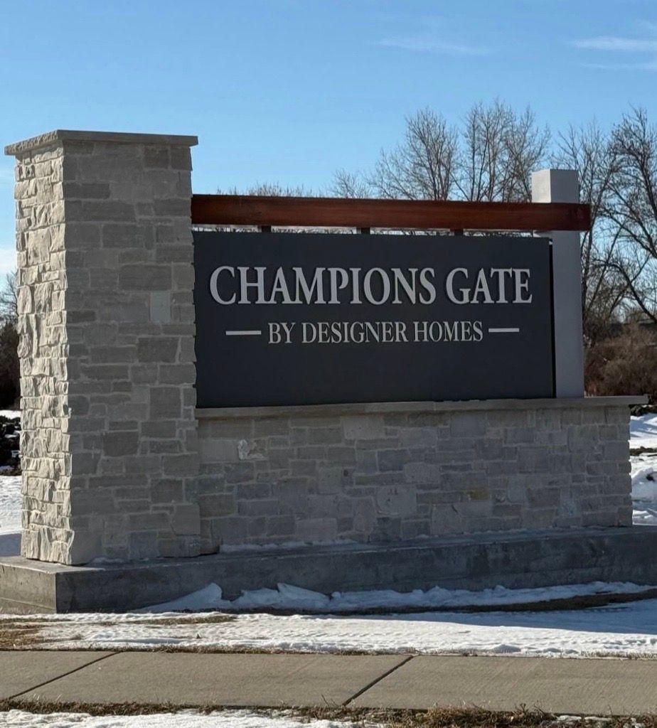 Stone entrance sign reading “Champions Gate” by Designer Homes under a blue sky, with snow on the ground