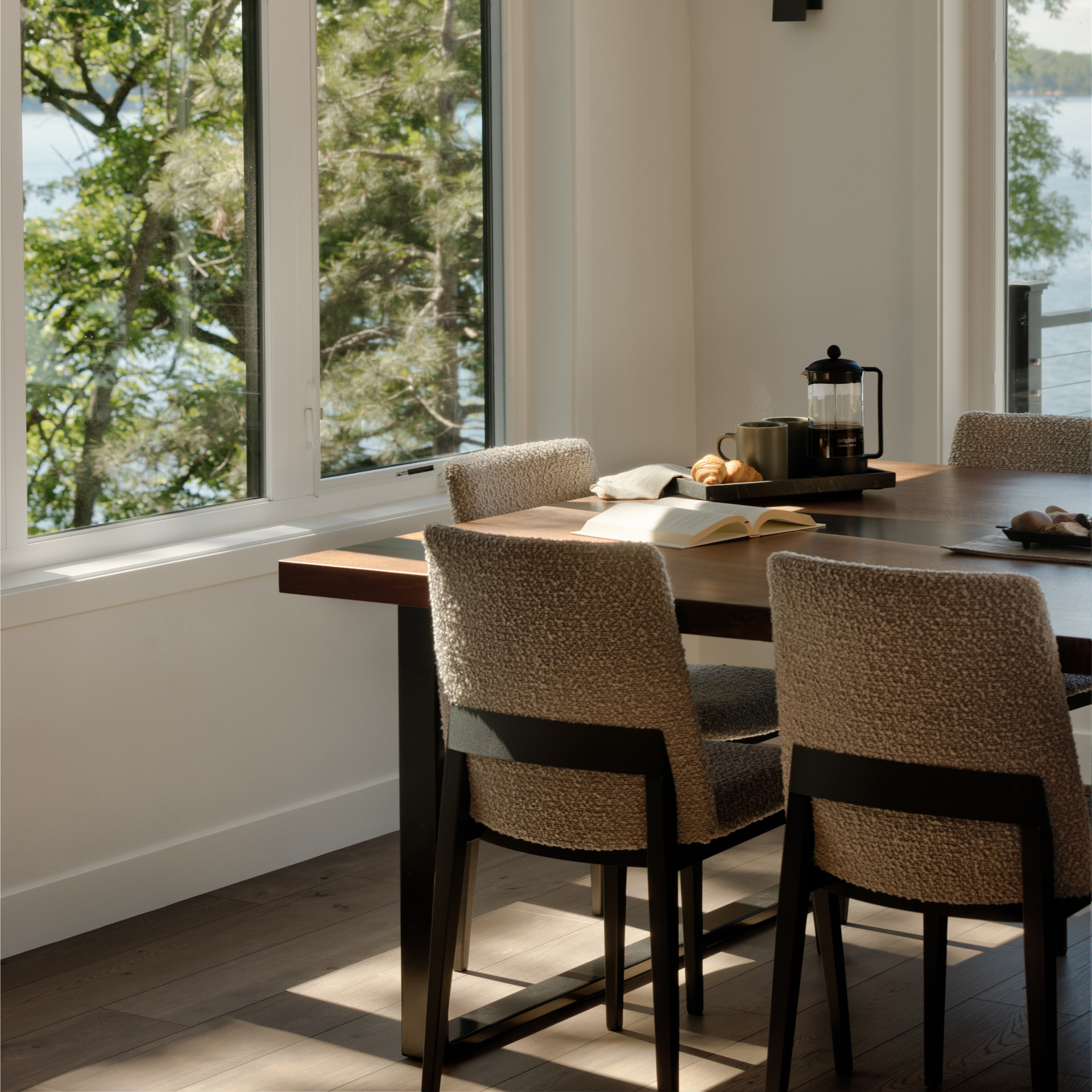 Sunlit dining room with a wooden table, upholstered chairs, and large windows overlooking trees