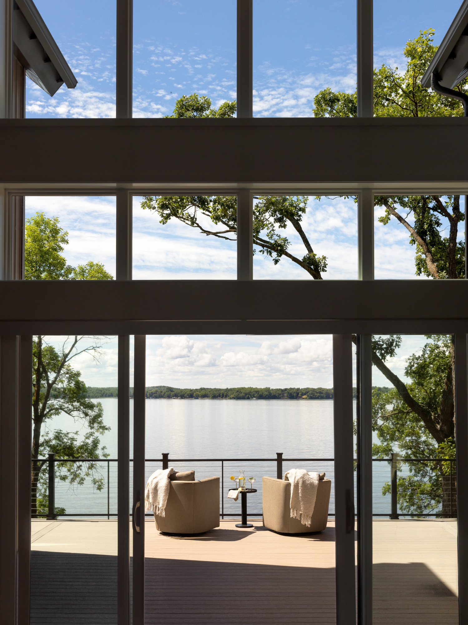 Modern patio with two beige chairs facing a lake, framed by large windows and greenery