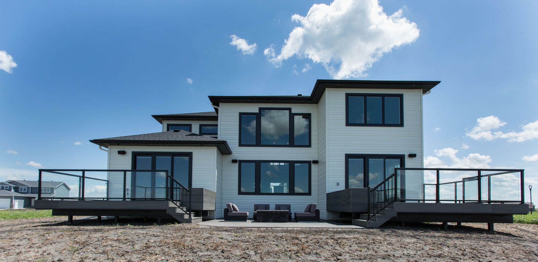 Back of a modern white house with black trim, two decks with glass railings, and a blue sky with clouds.