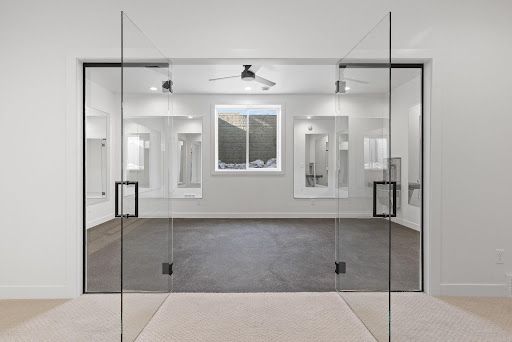 Glass double doors opening to a bright empty room with gray carpet and windows.