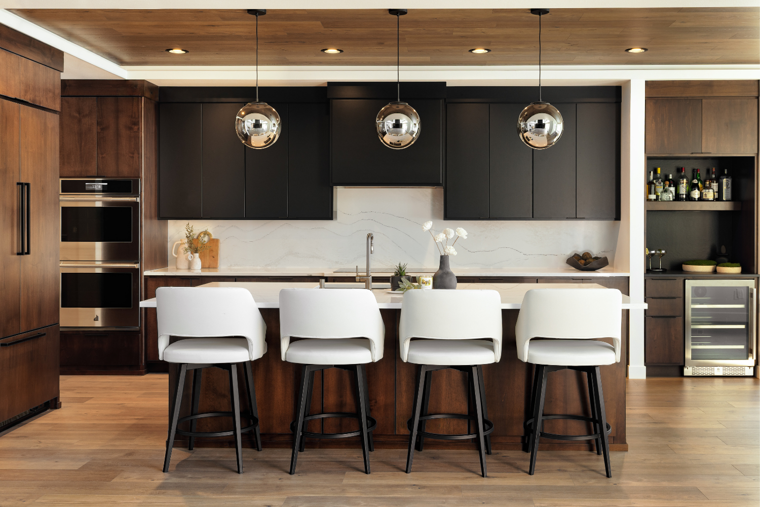 Modern kitchen with white island stools, dark cabinets, wood accents, and built-in appliances