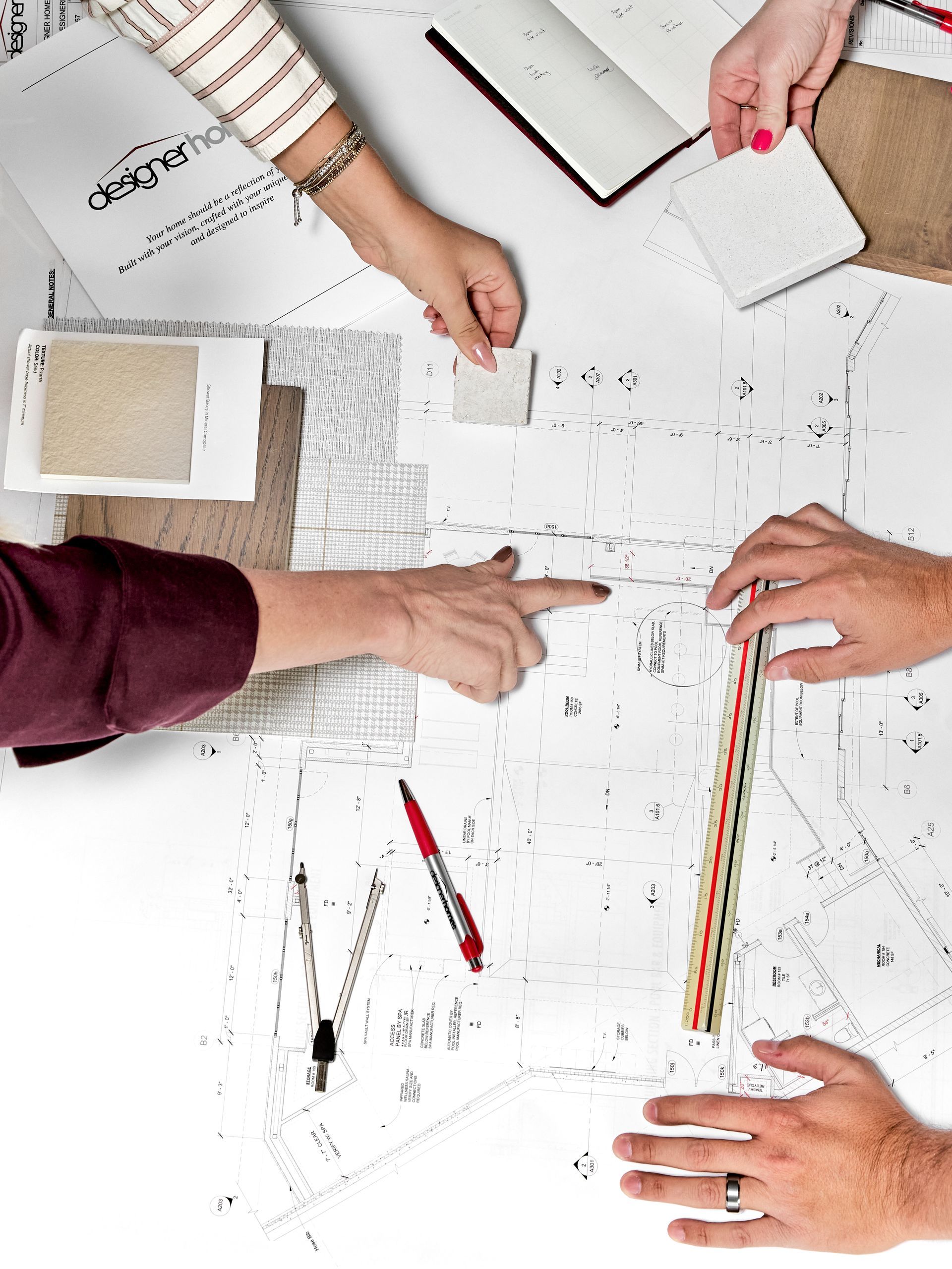 Hands collaborating over a white desk with sketches, rulers, pens, and color samples.
