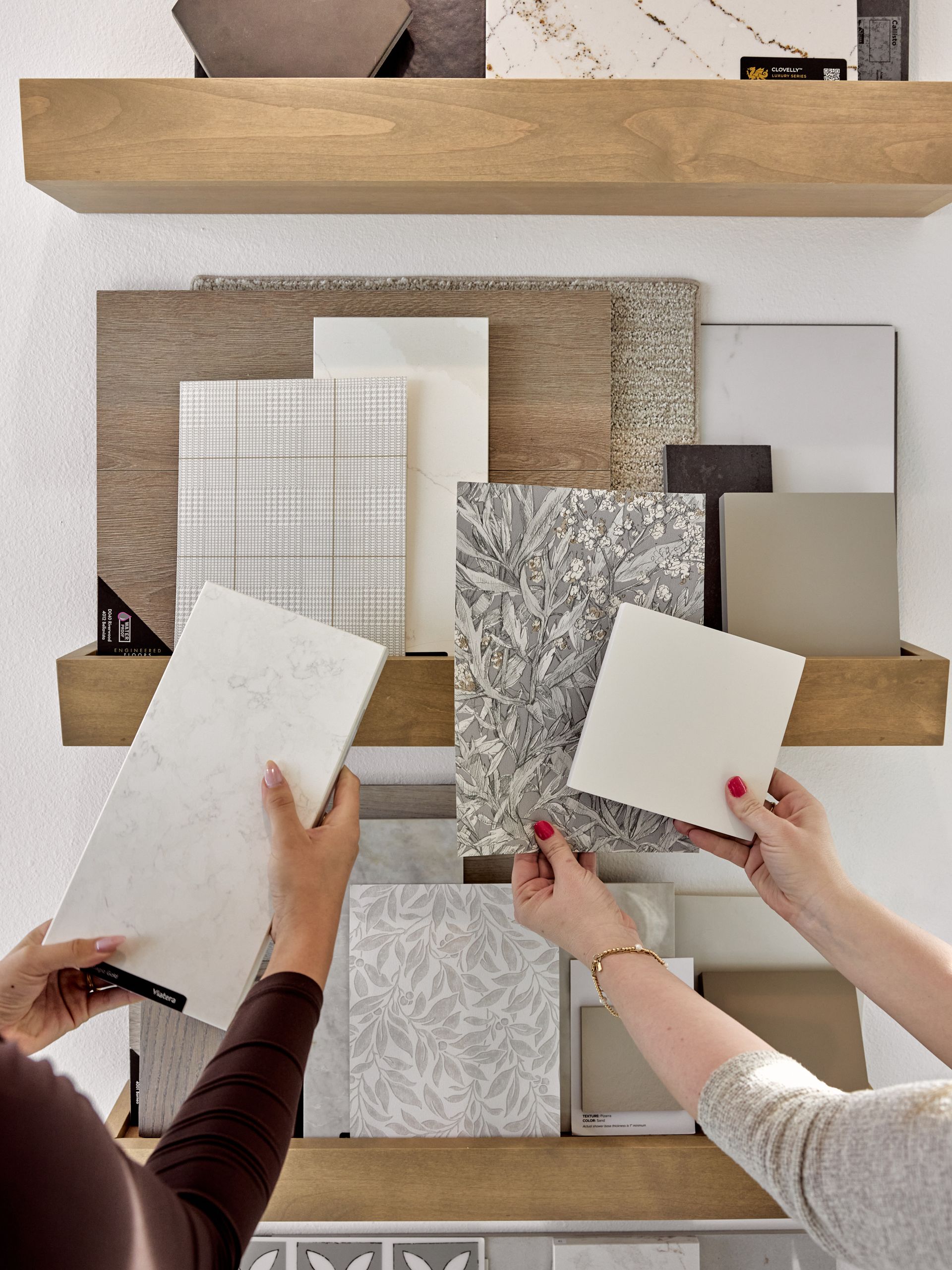 Hands comparing white paint swatches against a wall of neutral fabric samples in a showroom.