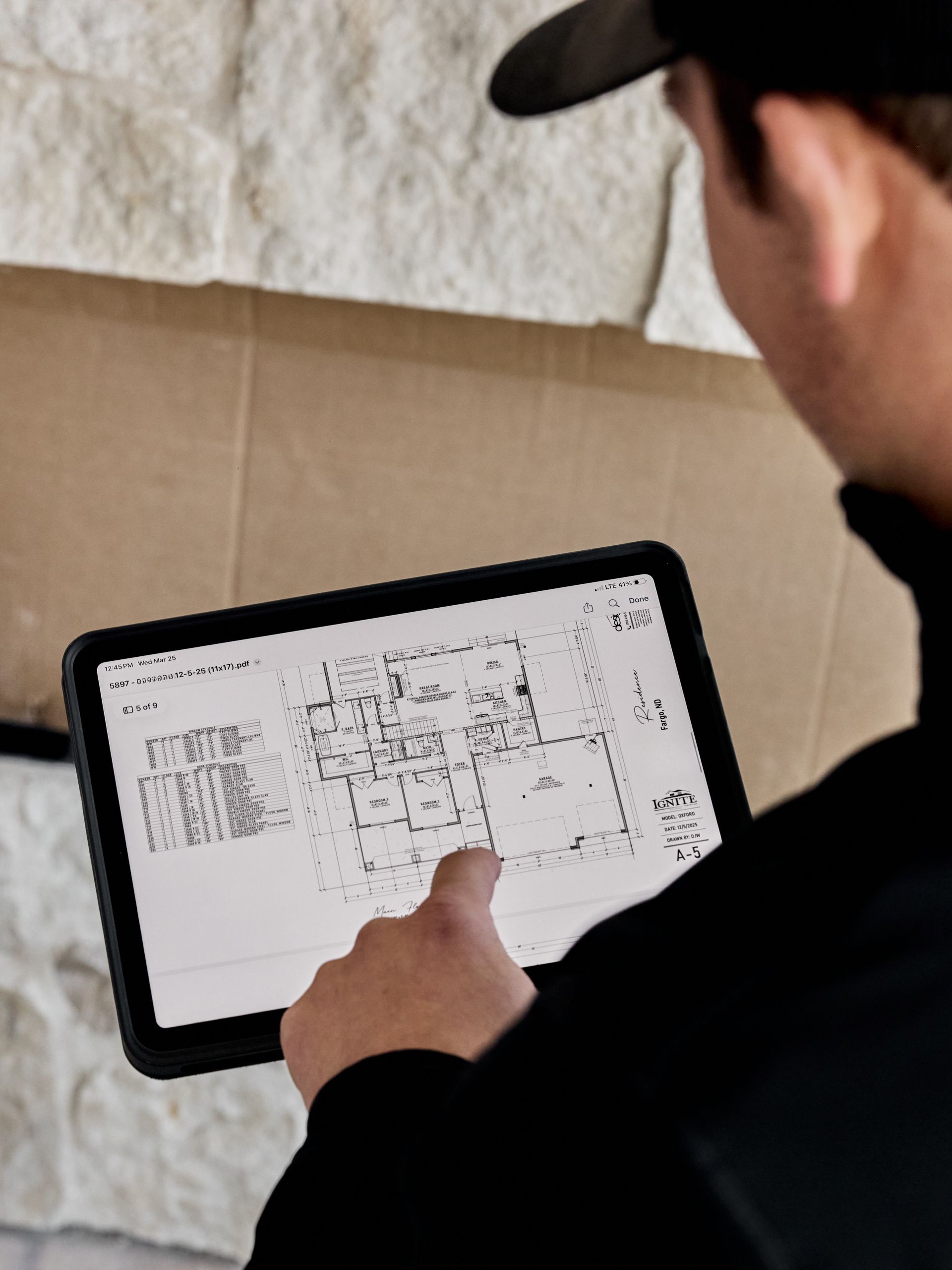 Person in a black cap pointing at a floor plan on a tablet in a work setting.
