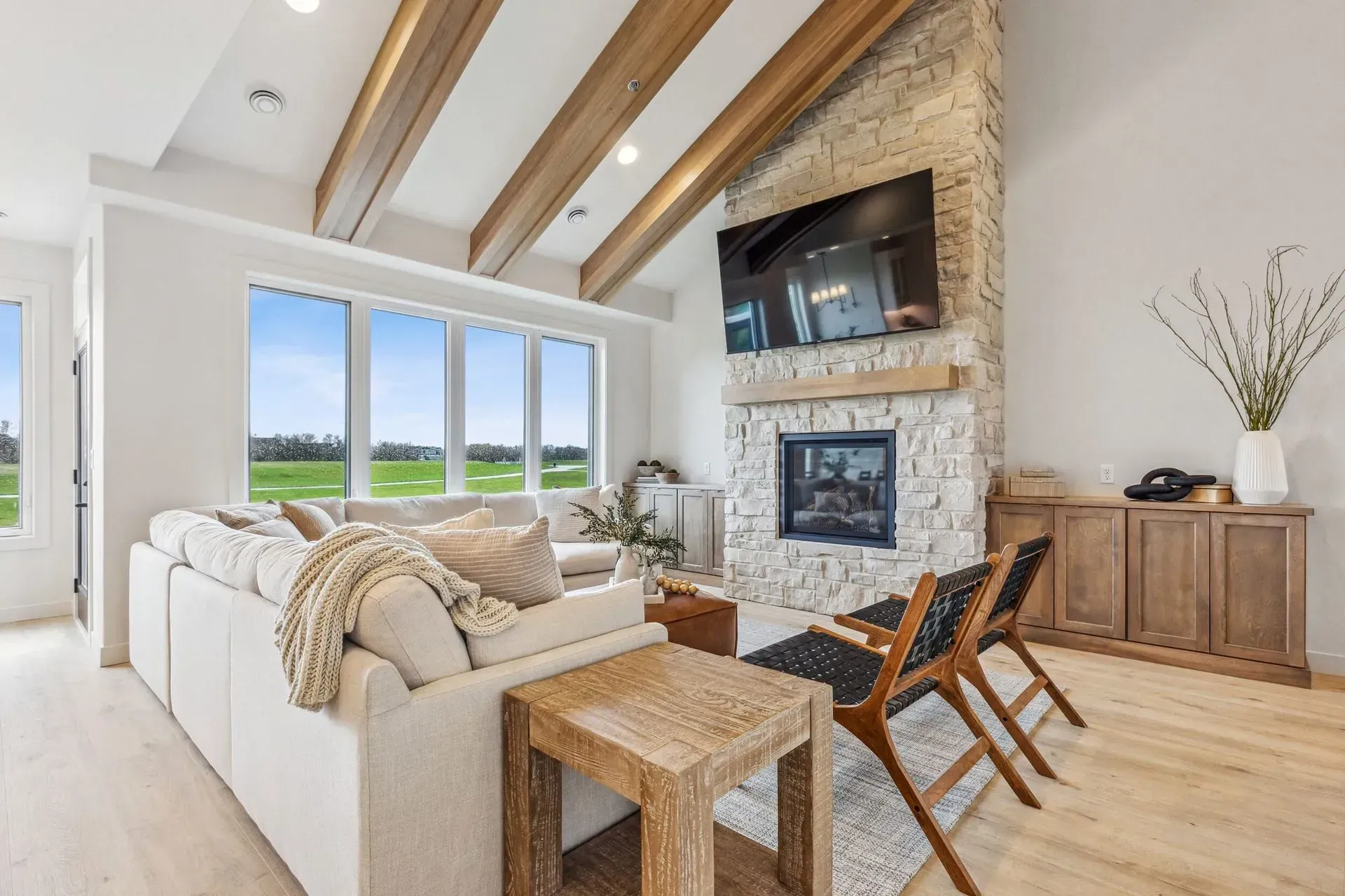 Bright living room with stone fireplace, white sofa, wood beams, and large windows overlooking fields