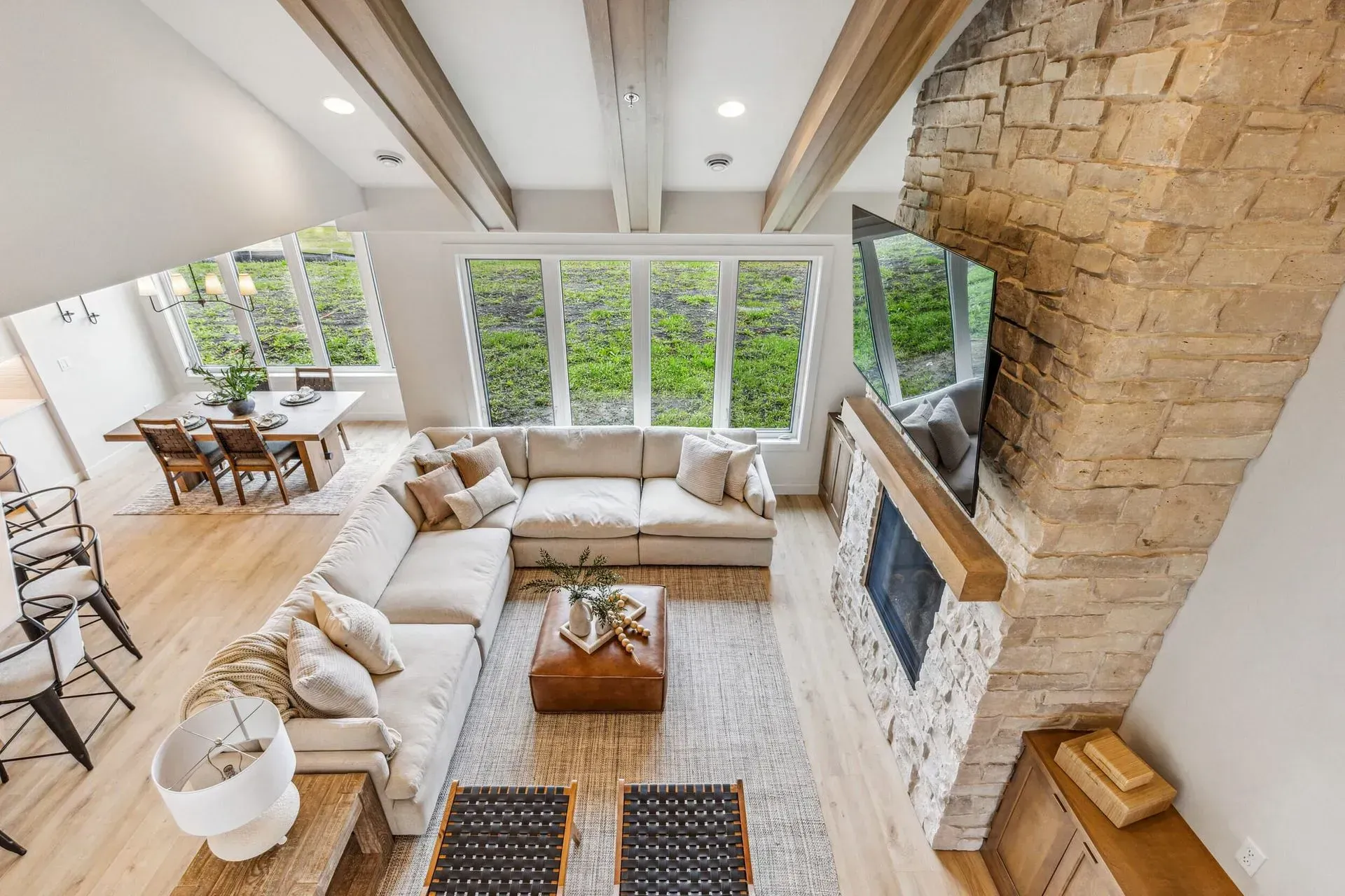 Bright modern living room with beige sofas, stone fireplace, wood beams, and large windows.