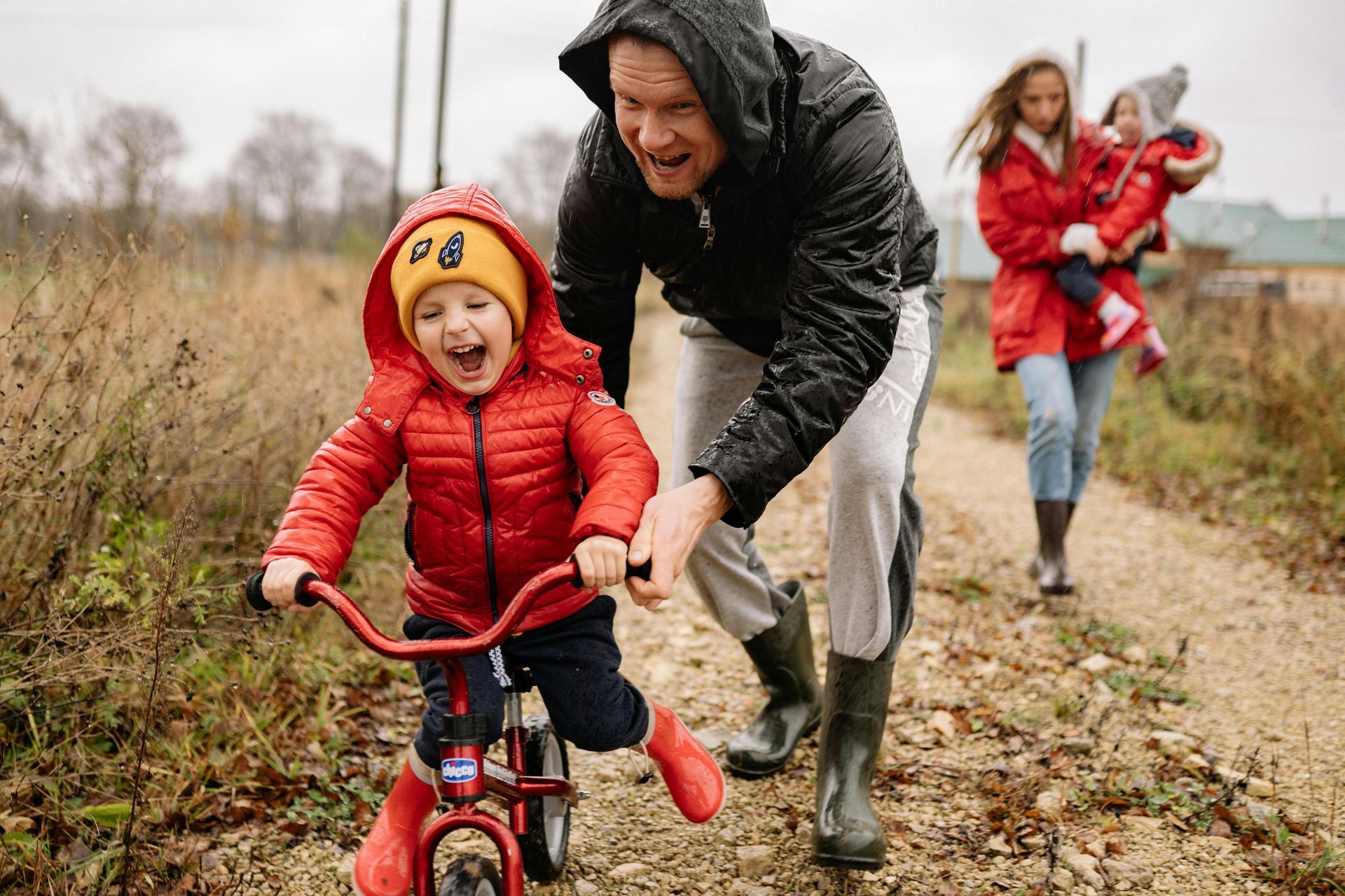 A smiling child on a red balance bike is pushed by an adult along a gravel path, with another person holding a toddler.