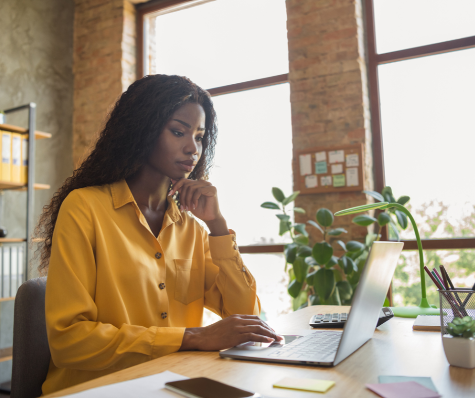 Woman in yellow shirt working on laptop at desk by window.
