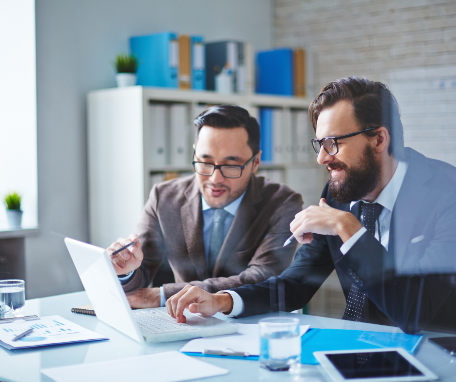 Two men in suits look at a laptop, one points, in an office.