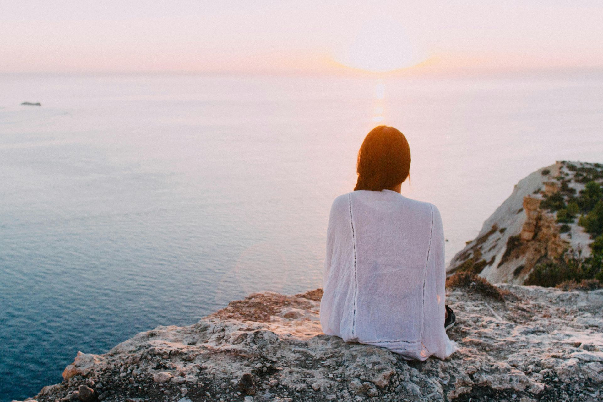 Woman in white shawl sits on cliff, looking at sunset over the ocean.