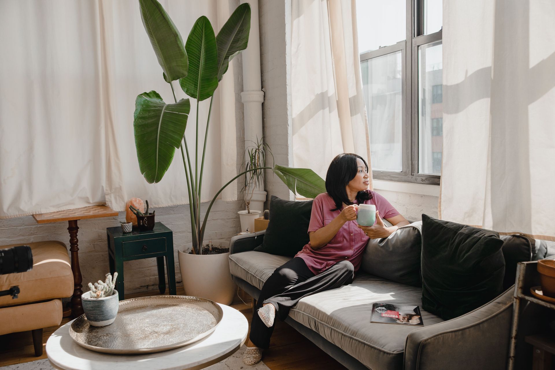 Person relaxing on a sofa by a bright window, holding a cup in a cozy living room with plants and curtains.