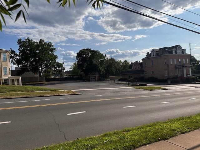 Quiet suburban street with a large house, trees, and cloudy sky across a wide road