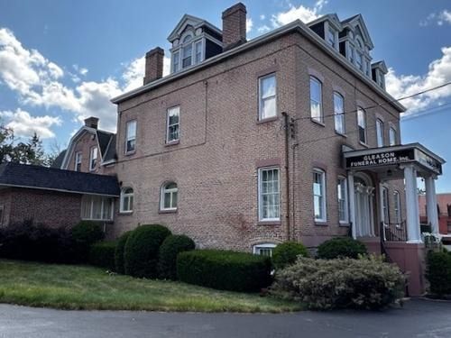 Brick building with a porch and dormer windows, surrounded by shrubs under a partly cloudy sky